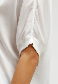 Hvid bomuldsbluse med puffede ærmer, samlet detalje ved skulderen og strikket manchet. Glat tekstur med subtil glans.