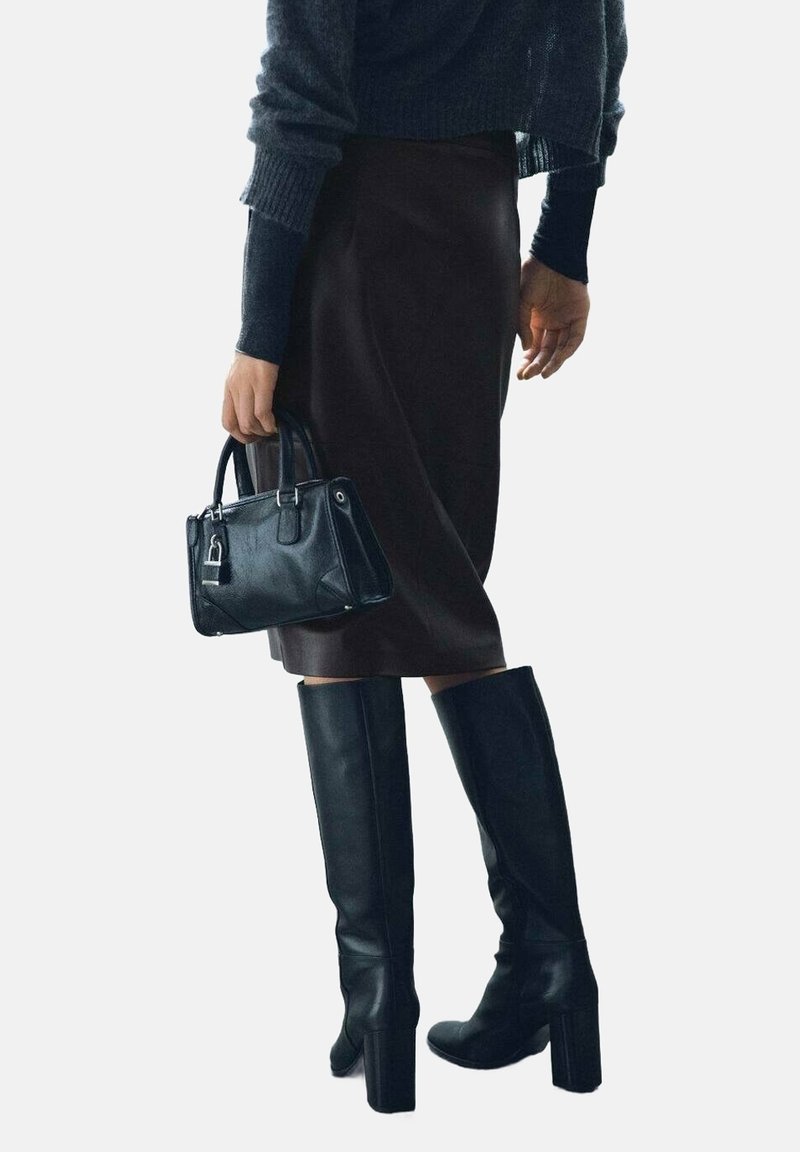 Personne portant des bottes noires montantes à talons et une jupe marron, tenant un petit sac à main noir à côté d’elle.
