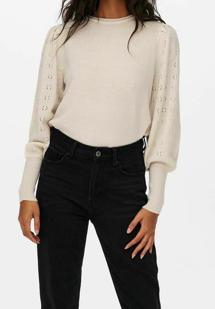 Femme portant un pull en maille beige à manches longues bouffantes avec motif œillet et un pantalon noir taille haute.