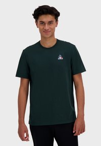 T-shirt en coton vert foncé avec un col rond et des manches courtes, orné d'un petit patch logo sur la poitrine.