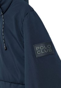 Navyblå vadderad jacka med en texturerad yta, utrustad med en svart gummibelagd "POLO CLUB" logotyp på ärmen och justerbara dragskor.