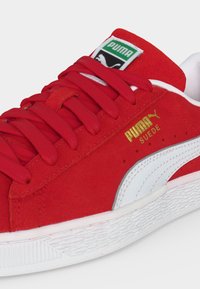 Κόκκινο σουέντ αθλητικό παπούτσι Puma με λευκή ρίγα, χρυσό λογότυπο, κόκκινα κορδόνια και πράσινη ετικέτα Puma στη γλώσσα.