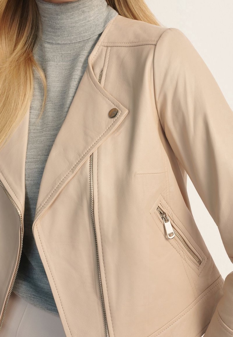 Femme portant une veste en cuir beige par-dessus un pull à col roulé gris clair, montrant les détails de la fermeture éclair et des boutons-pression de la veste.