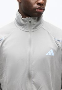 Άνδρας που φοράει αθλητικό μπουφάν με φερμουάρ σε ανοιχτό γκρι χρώμα, με ψηλό γιακά και μικρό μπλε λογότυπο Adidas στο στήθος, στραμμένος ελαφρώς προς τα δεξιά.