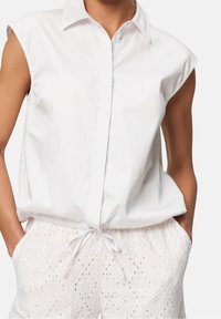 Blouse blanche à col, manches courtes, fermeture à boutons, associée à un short blanc texturé avec un motif œillet géométrique et un cordon de serrage.