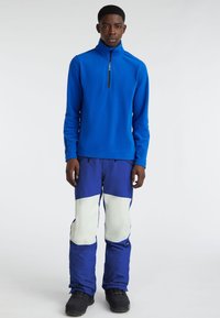 Blue fleece half-zip pullover with a standing collar, paired with blue snow pants featuring white panel accents and black boots.