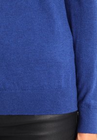 Maglione blu a maniche lunghe con polsini a costine indossato sopra pantaloni in pelle nera, che mostra il torso e il braccio destro di una persona.