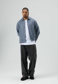 Blauw patroon overhemd over een wit T-shirt, gecombineerd met loszittende donkergrijze broek en zwarte schoenen met roze veters.