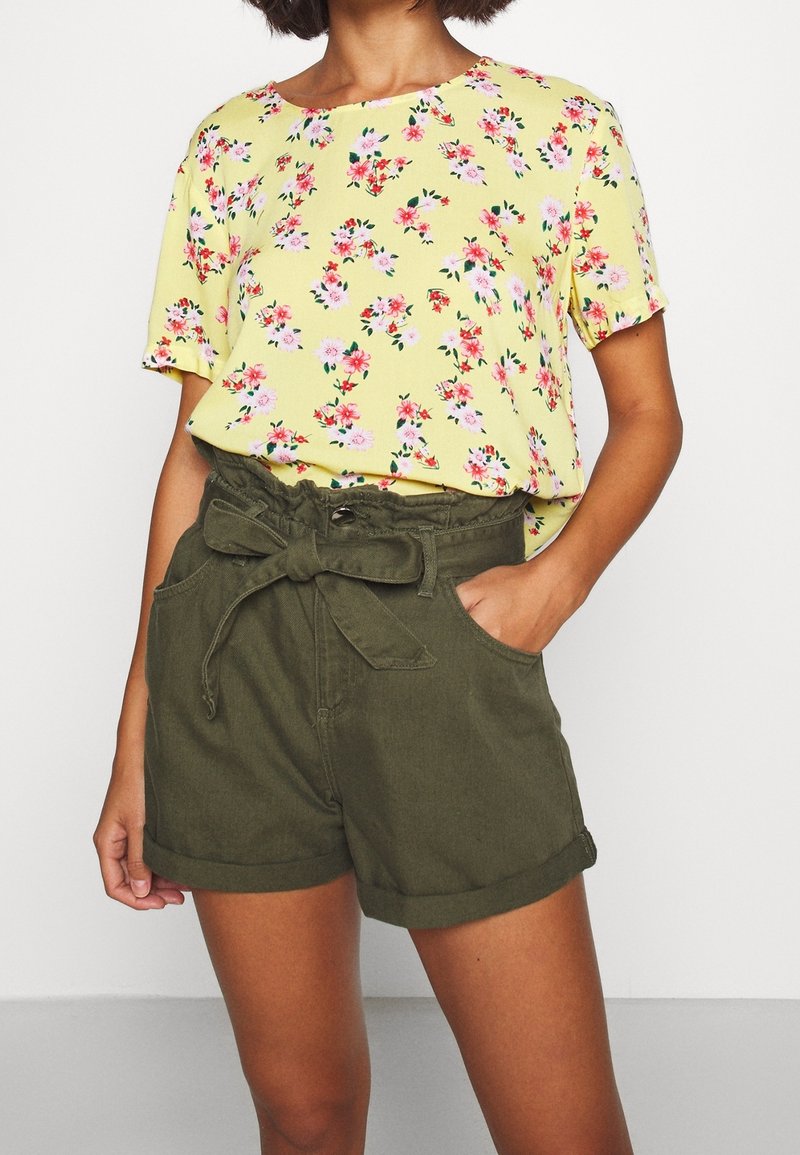 Žlutá květinová blůza s krátkým rukávem v kombinaci s olivově zelenými šortkami s vysokým pasem, které mají svazovací pásek a boční kapsy.