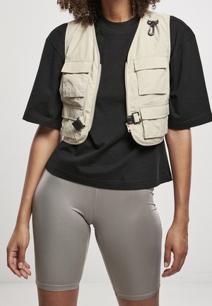 Beige mouwloos vest met meerdere zakken, ritssluiting en zwart beslag, gedragen over een zwart t-shirt en grijze fietsshorts.