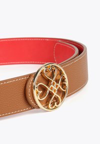 En läderbälte i tan med en slät yta och en gyllene rund spänne med en intrikat hjärt-design. Inuti är det röd. Vita söm detaljer.