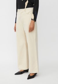 Persona che indossa pantaloni beige a gamba larga, scarpe nere a punta e una maglia nera a maniche lunghe con pattern beige e navy, in piedi davanti a uno sfondo bianco.