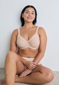 Beige bra with wide straps, smooth texture, and seam details. Paired with matching light beige panties, sitting on a neutral-toned surface.