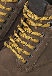 Des bottes en cuir marron avec des lacets jaunes et noirs à motif, des œillets en métal, et une surface texturée avec des détails de couture.