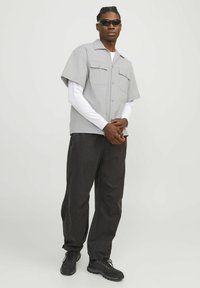 Graues Kurzarmhemd mit zwei Brusttaschen über einem weißen Langarmshirt, kombiniert mit weiten schwarzen Hosen und schwarzen Turnschuhen.