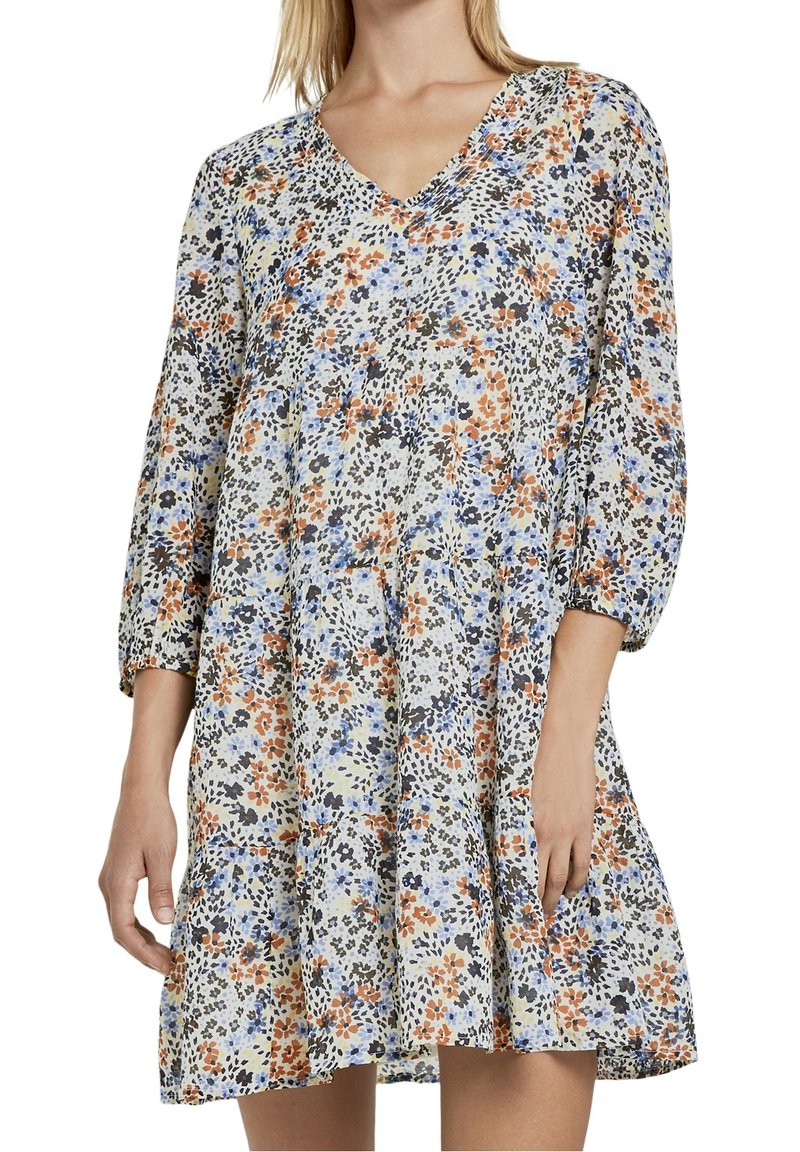 Femme portant une robe ample à motif floral avec manches 3/4 et col en V, présentant des fleurs bleues, orange, noires et jaunes sur tissu blanc.