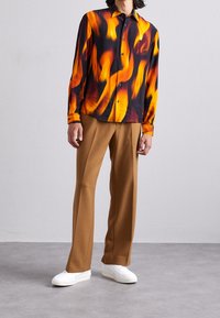 Košile s dlouhým rukávem s černým základem a vzory oranžových a žlutých plamenů, zkombinována s hnědými kalhotami a bílými teniskami.