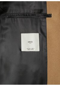 Bruine stof met een gladde textuur aan één kant; zwarte voering met een wit label waarop "MNG MAN" en maatdetails staan.