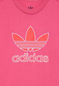 Rozā kokvilnas t-krekls ar baltu kontūru "adidas" logo un trim lapu formas svītrām krūtīs. Klasiska apļa kakla forma.