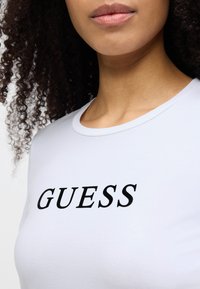 White short-sleeve t-shirt with a round neckline featuring black "GUESS" text at the front. Smooth texture, form-fitting design.