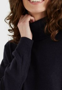 Maglione nero a collo alto in tessuto morbido e testurizzato. Presenta polsini a costine e una vestibilità rilassata, con un primo piano di una mano vicino al collo.