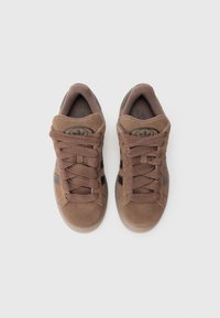 Brown suede sneakers with textured uppers, round laces, and dark accents. Features a flat sole and padded collar for comfort.