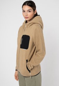 Beige Fleece-Hoodie mit einer schwarzen Reißverschlusstasche auf der Brust, Kängurutaschen und einer Kapuze mit Kordelzug; besitzt eine weiche Textur und eine lockere Passform.