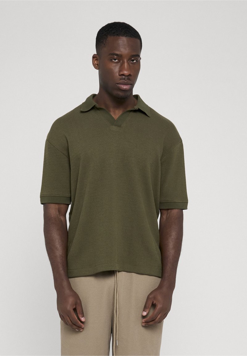 Jeune homme debout, portant un polo à manches courtes texturé vert olive foncé et un pantalon beige à cordon, sur un fond clair uni.