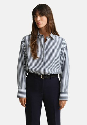 Vrouw met lang bruin haar, gekleed in een blauw-wit gestreept overhemd met verticale strepen, ingestopt in donkerblauwe broek met een zwarte riem.