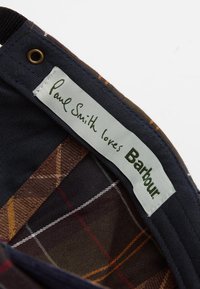 Σήμα Paul Smith x Barbour σε ένδυμα που αναδεικνύει καρό ύφασμα σε καφέ και ναυτικό μπλε με πορτοκαλί λεπτομέρειες; διαθέτει ραμμένο ετικέτα.