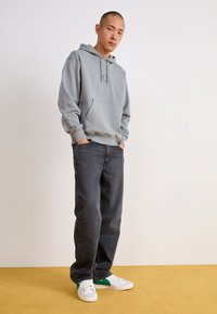 Sudadera gris con bolsillo frontal y cordón, combinada con jeans gris oscuro y zapatillas blancas con detalles verdes sobre un suelo amarillo.
