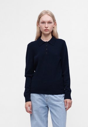 Jeune femme aux longs cheveux blonds porte un pull polo noir à manches longues et un jean bleu clair, debout devant un fond blanc uni.