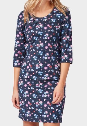 Femme portant une robe bleu marine avec des motifs floraux rose, blanc et bleu, à manches trois-quarts, debout devant un fond blanc.