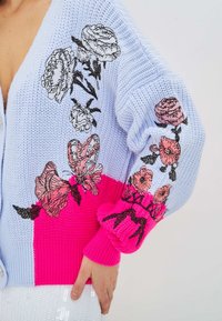 Knit cardigan with a light blue body, bright pink bottom, and floral embroidery in pink and black on sleeves and front.