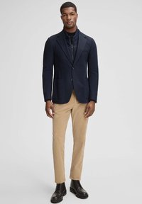 Granatowa marynarka o wyraźnej, strukturalnej formie, z dwoma przednimi kieszeniami i czarnymi guzikami, zestawiona z khaki spodniami i czarnymi butami sznurowanymi.