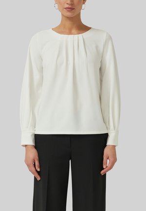Vrouw draagt een witte blouse met lange mouwen en voorplooien, en zwarte op maat gemaakte broek, staand tegen een effen grijze achtergrond.