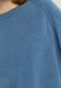 Niebieski sweter o miękkiej fakturze. Posiada okrągły dekolt i ribbowane krawędzie. Materiał wydaje się lekki i gładki.