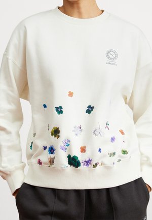 Biała bluza z małymi, rozrzuconymi kolorowymi kwiatowymi wzorami i logo LIBERTY na górnej lewej części klatki piersiowej, noszona z ciemnymi spodniami.