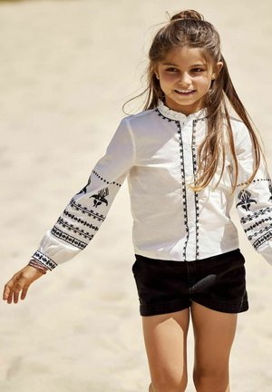 Chemisier blanc avec des motifs brodés noirs sur les manches et le devant, col volanté, associé à un short noir. Tissu doux, coupe décontractée.