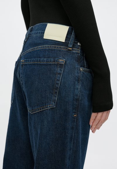 Sötétkék, lazán szabott farmernadrág, sima textúrával, kontrasztos narancssárga varrással és világos színű bőr címkével a hátsó részén.
