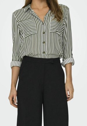 Femme portant une blouse à rayures verticales vertes et blanches à manches longues avec poches avant et pantalon noir taille haute, manches retroussées.