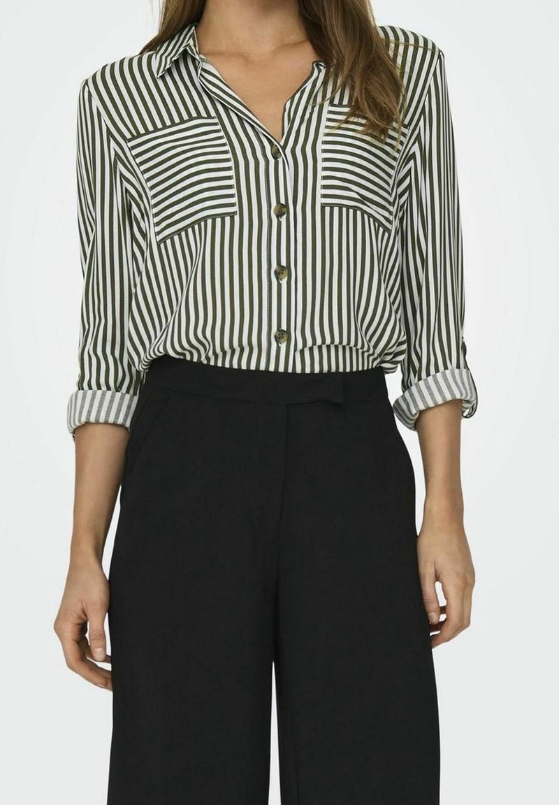 Femme portant une blouse à rayures verticales vertes et blanches à manches longues avec poches avant et pantalon noir taille haute, manches retroussées.
