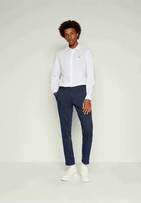 Vit långärmad skjorta med knappar, marinblå byxor och vita sneakers. Inkluderar en liten logotyp på skjortan. Slim fit-design.