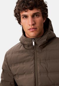 Chaqueta acolchada marrón con capucha, que presenta un cierre de cremallera, costuras horizontales y una superficie texturizada. Logo visible en el material.