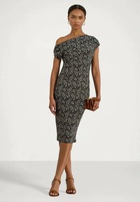 Mulher vestida com um vestido midi de uma só alça em padrão paisley preto e branco, segurando uma carteira castanha e usando sapatos de salto com tiras no tornozelo castanhos, de pé contra um fundo branco.