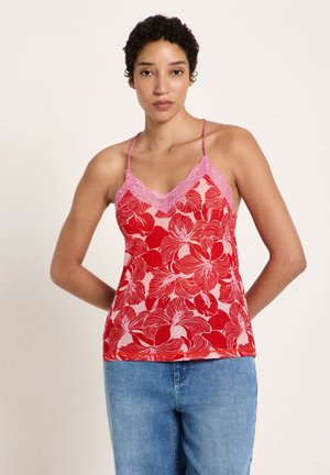 Woman with short curly hair wears red floral camisole with pink lace trim and blue jeans, standing with hands behind back.