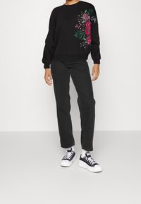 Sudadera negra con bordado floral en rosa y verde en el lado izquierdo, combinada con jeans negros de pierna recta y zapatillas en blanco y negro.