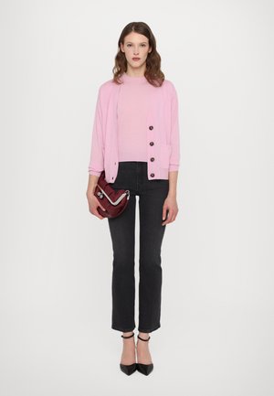 Cardigan rosa con bottoni sopra una maglietta rosa chiaro, abbinato a jeans neri a 3/4 e décolleté neri. La modella tiene una borsa bordeaux.
