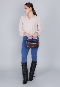 Kvinna som står mot en grå bakgrund, iklädd en beige blus, blå jeans, svarta knähöga stövlar och håller en mörkbrun handväska med guldhandtag.