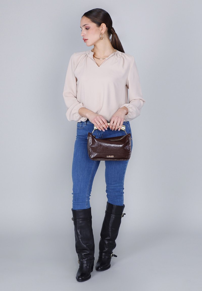 Kvinna som står mot en grå bakgrund, iklädd en beige blus, blå jeans, svarta knähöga stövlar och håller en mörkbrun handväska med guldhandtag.